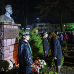 Pomnik Piłsudskiego nocą, otoczony zielenią. Grupa ludzi w tle, światła i wieńce. Uroczystość upamiętniająca.