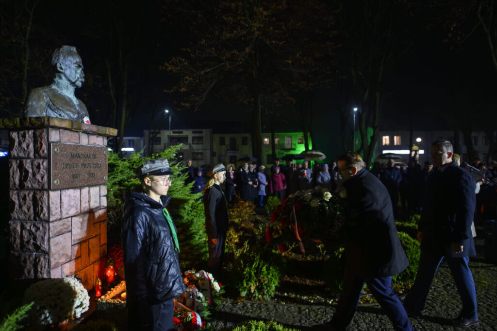 Pomnik Marszałka Piłsudskiego otoczony wianuszkiem. Tło nocne, podświetlone na zielono budynki i zgromadzeni ludzie. Osoby w historycznych mundurach składają hołd.