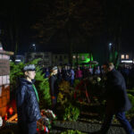 Pomnik Marszałka Piłsudskiego otoczony wianuszkiem. Tło nocne, podświetlone na zielono budynki i zgromadzeni ludzie. Osoby w historycznych mundurach składają hołd.