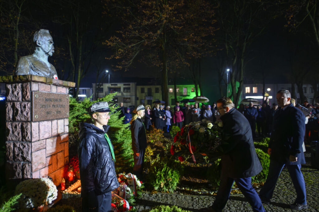 Pomnik z popiersiem w parku nocą, w blasku reflektorów. Wokół ludzie w odświętnych strojach składają kwiaty. Tło to drzewa i oświetlone budynki.