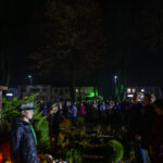 Pomnik na tle tłumu w parku nocą. Osoby w mundurach stoją po lewej. Otoczenie oświetlone jest zielonym i niebieskim światłem.