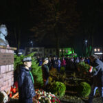 Popiersie marszałka na postumencie z cegły otoczonego wieńcami. Nocna sceneria z ludźmi i zielonymi krzewami w tle. Oświetlone budynki i drzewa.