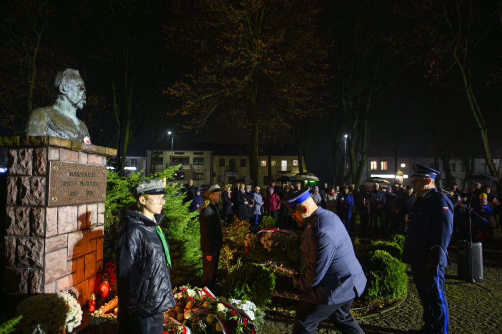 Pomnik Piłsudskiego otoczony kwiatami i ludźmi w mundurach. Nocne tło z drzewami, delikatne oświetlenie. Ludzie zgromadzeni wokół.