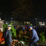 Pomnik Piłsudskiego otoczony kwiatami i ludźmi w mundurach. Nocne tło z drzewami, delikatne oświetlenie. Ludzie zgromadzeni wokół.