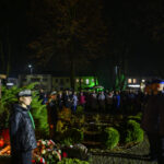 Pomnik Piłsudskiego z boku, ciemna noc. Ludzie wokół, światła z tyłu. Zieleń i znicze pod pomnikiem.