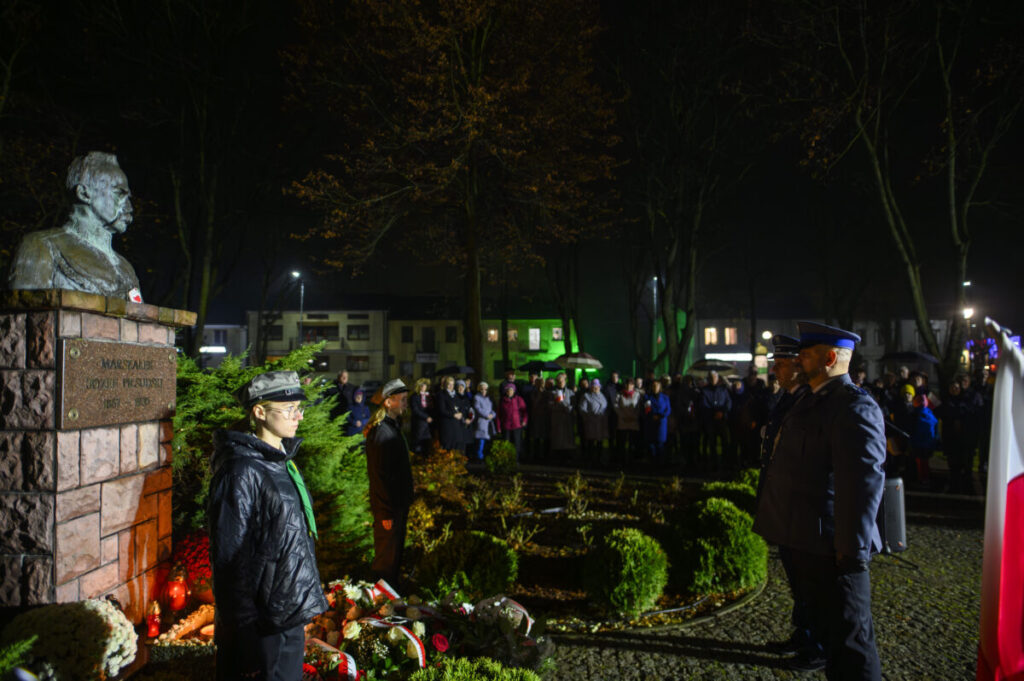 Posąg z brązu na kamiennym cokole w parku nocą. Przed pomnikiem stoją ludzie w mundurach. W tle drzewa i tłum ludzi.