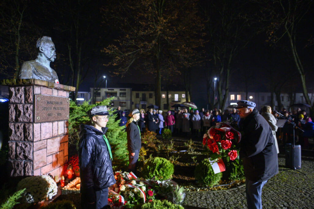 Pomnik Piłsudskiego oświetlony nocą, otoczony przez ludzi z biało-czerwonymi kwiatami. Tło to ciemne niebo i drzewa. Wydarzenie upamiętnia ważną osobę.