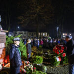 Pomnik Piłsudskiego oświetlony nocą, otoczony przez ludzi z biało-czerwonymi kwiatami. Tło to ciemne niebo i drzewa. Wydarzenie upamiętnia ważną osobę.