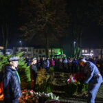 Biały pomnik na tle nocnym, otoczony ludźmi w mundurach. Uczestnicy składają kwiaty. Tło z wysokimi drzewami i budynkami.