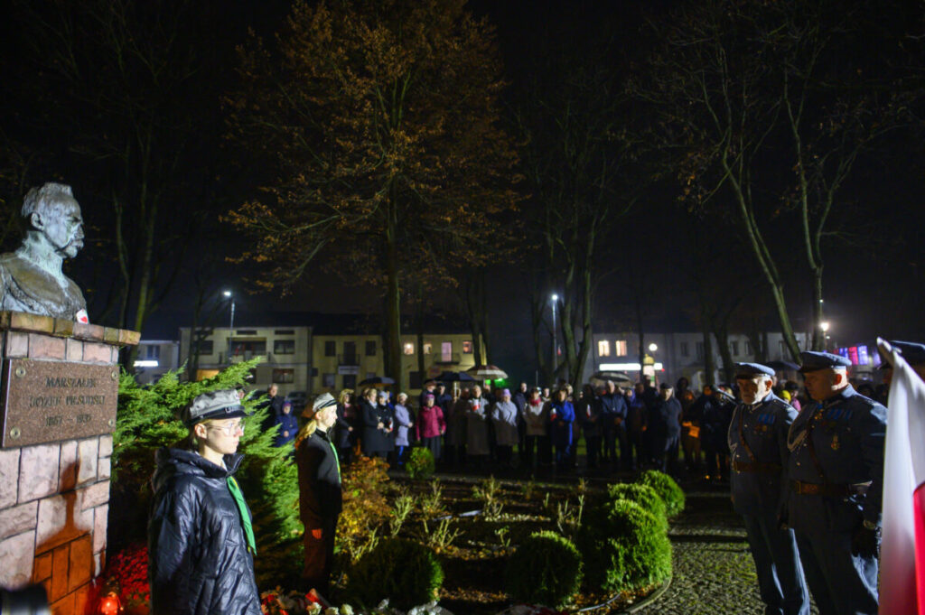 Pomnik Marszałka Piłsudskiego nocą otoczony przez ludzi. Tło z drzewami, w oddali widoczne budynki. Oświetlenie białe i żółte.
