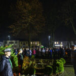 Pomnik Marszałka Piłsudskiego nocą otoczony przez ludzi. Tło z drzewami, w oddali widoczne budynki. Oświetlenie białe i żółte.