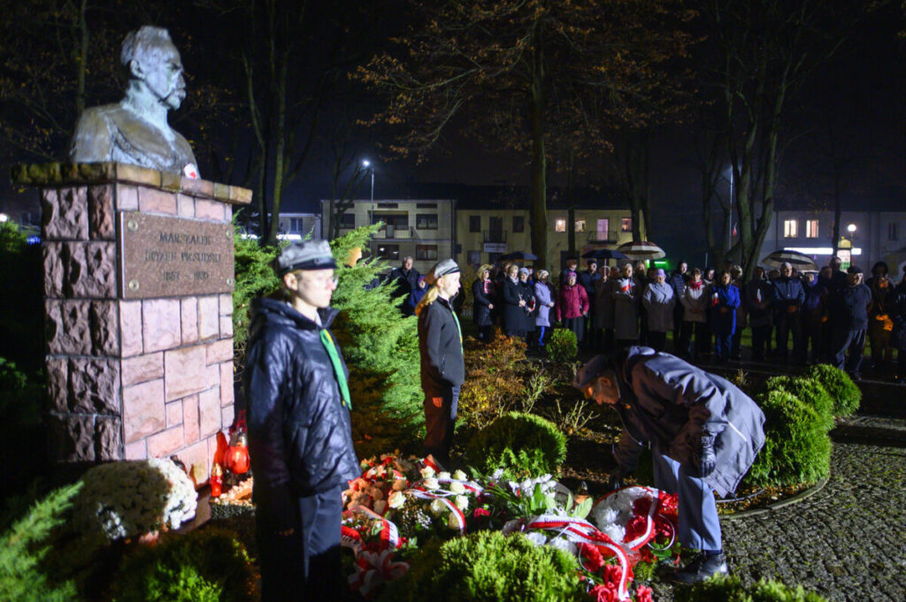Pomnik z popiersiem mężczyzny stoi w parku nocą. Wokół są ludzie, żołnierze i kwiaty w biało-czerwonych barwach. Za tłem widać budynki.