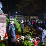 Pomnik z popiersiem mężczyzny stoi w parku nocą. Wokół są ludzie, żołnierze i kwiaty w biało-czerwonych barwach. Za tłem widać budynki.