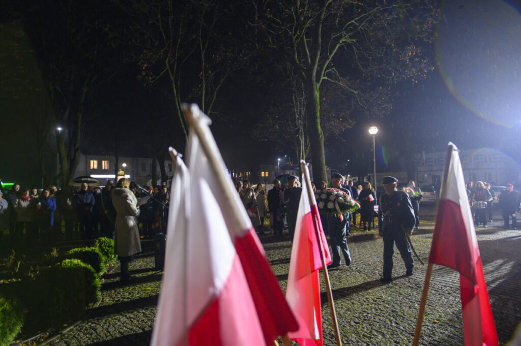 Ludzie zebrani na nocnej ceremonii z flagami Polski. Ciemne drzewa w tle, oświetlone latarniami. Postać z wieńcem kwiatów.