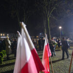 Ludzie zebrani na nocnej ceremonii z flagami Polski. Ciemne drzewa w tle, oświetlone latarniami. Postać z wieńcem kwiatów.