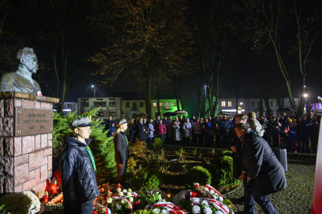 Pomnik w parku podczas nocnej uroczystości. Ludzie zgromadzeni wokół, z kwiatami i flagami. Oświetlenie w odcieniach zieleni.