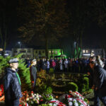 Pomnik w parku podczas nocnej uroczystości. Ludzie zgromadzeni wokół, z kwiatami i flagami. Oświetlenie w odcieniach zieleni.