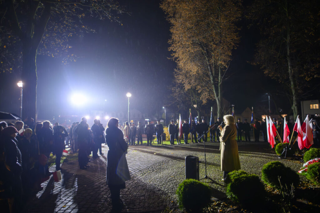 Wieczorna uroczystość z przemawiającą osobą i publicznością pod drzewami. Na pierwszym planie flagi biało-czerwone. Światła lamp oświetlają miejsce.