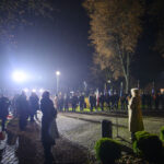 Wieczorna uroczystość z przemawiającą osobą i publicznością pod drzewami. Na pierwszym planie flagi biało-czerwone. Światła lamp oświetlają miejsce.