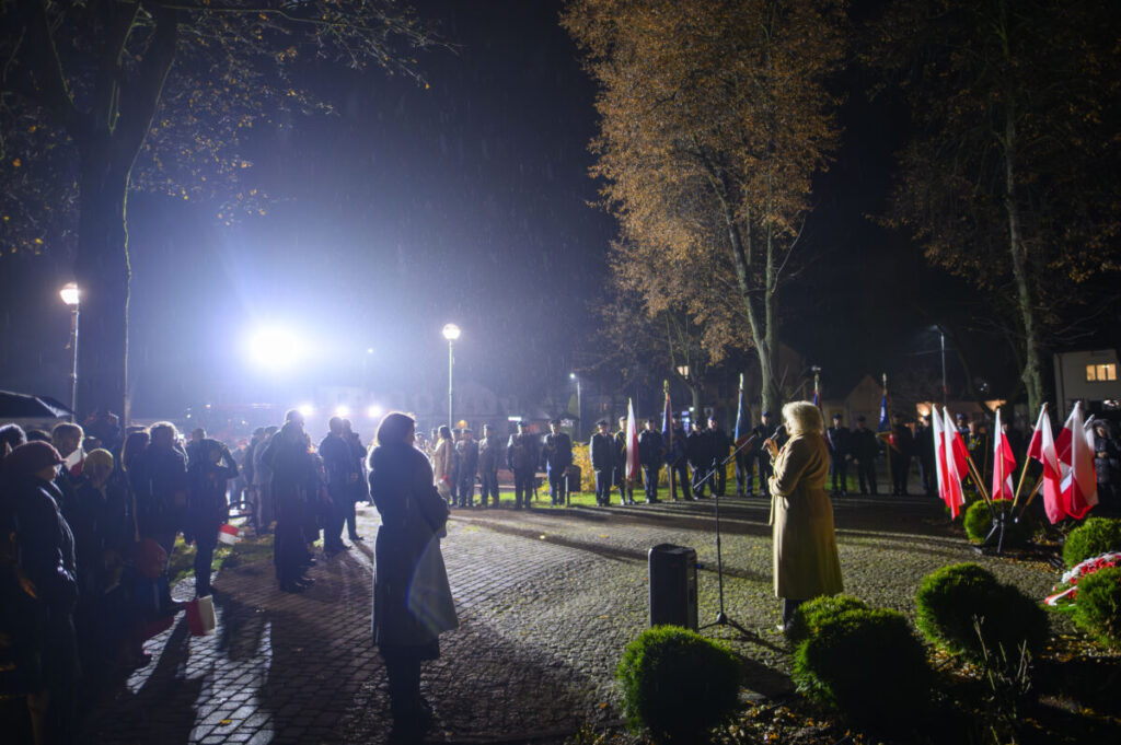 Nocna uroczystość na zewnątrz z przemawiającą osobą w jasnym płaszczu. W tle ludzie i polskie flagi w świetle lamp. Drzewa bez liści otaczają plac.