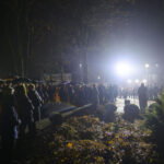 Nocna uroczystość; tłum ludzi pod parasolami. Silne światło w tle, okolica parku; jesienne liście na ziemi.