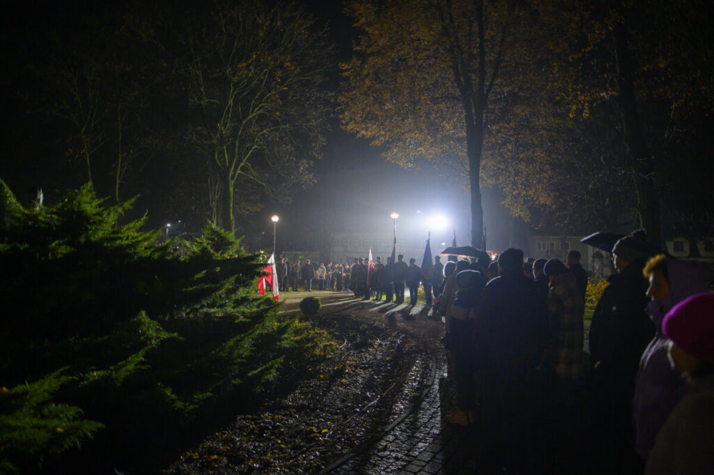 Nocna scena z tłumem ludzi stojących wzdłuż oświetlonej ścieżki. W tle drzewa, światła, jesienne liście. Flaga Polski widoczna po lewej.