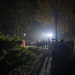 Nocna scena z tłumem ludzi stojących wzdłuż oświetlonej ścieżki. W tle drzewa, światła, jesienne liście. Flaga Polski widoczna po lewej.