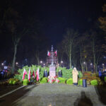 Pomnik otoczony zielenią z polskimi flagami na tle nocnego parku. Ludzie stoją wokół, słuchając przemówienia. Ciepłe światła oświetlają scenę.