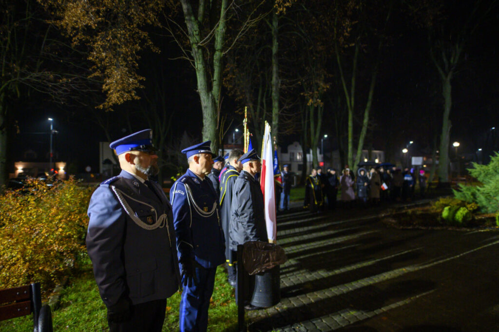Policjanci w niebieskich mundurach stoją w rzędzie nocą. W tle oświetlony park i ludzie z flagami. Obok rośliny i latarnie.