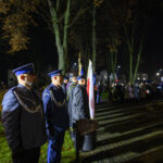 Policjanci w niebieskich mundurach stoją w rzędzie nocą. W tle oświetlony park i ludzie z flagami. Obok rośliny i latarnie.