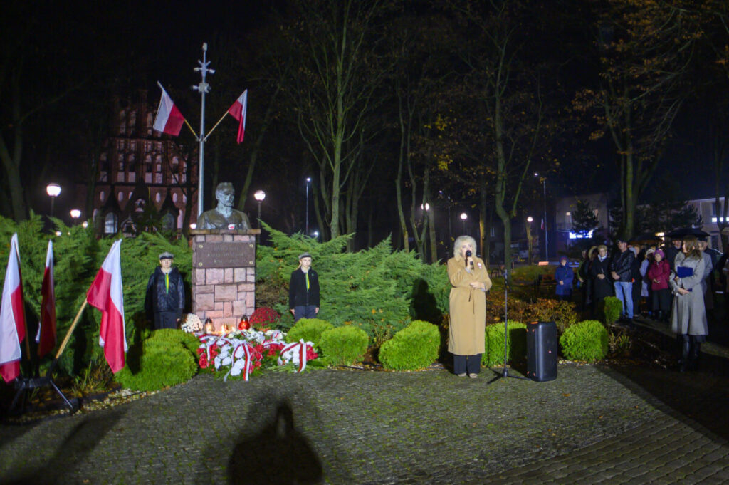 Na nocnym zdjęciu widoczny jest pomnik z popiersiem na tle drzew, flagi Polski i grupa ludzi. Obok stoi osoba z mikrofonem, a w tle znajdują się oświetlone budynki. Dominuje ciemne, zielono-brązowe tło.