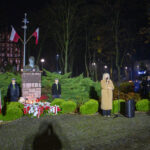 Na nocnym zdjęciu widoczny jest pomnik z popiersiem na tle drzew, flagi Polski i grupa ludzi. Obok stoi osoba z mikrofonem, a w tle znajdują się oświetlone budynki. Dominuje ciemne, zielono-brązowe tło.