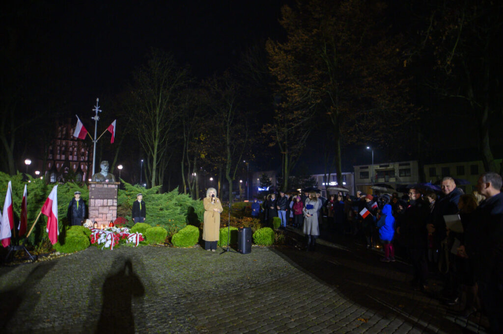 Wieczorna uroczystość na placu z pomnikiem i polskimi flagami. Kobieta w płaszczu przemawia, otoczona przez tłum. Tło stanowią drzewa i budynek.