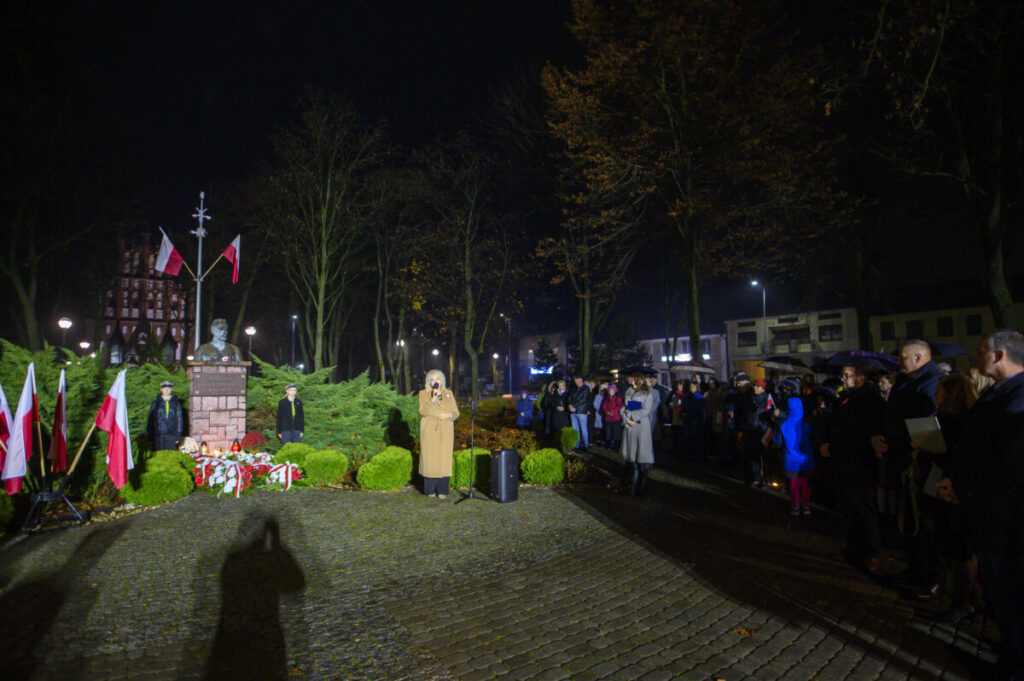 Nocna uroczystość przy pomniku z flagami i kwiatami. Kobieta przemawia, ludzie zgromadzeni obok. Drzewa w tle i ceglana droga.