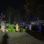 Nocna uroczystość przy pomniku z flagami i kwiatami. Kobieta przemawia, ludzie zgromadzeni obok. Drzewa w tle i ceglana droga.