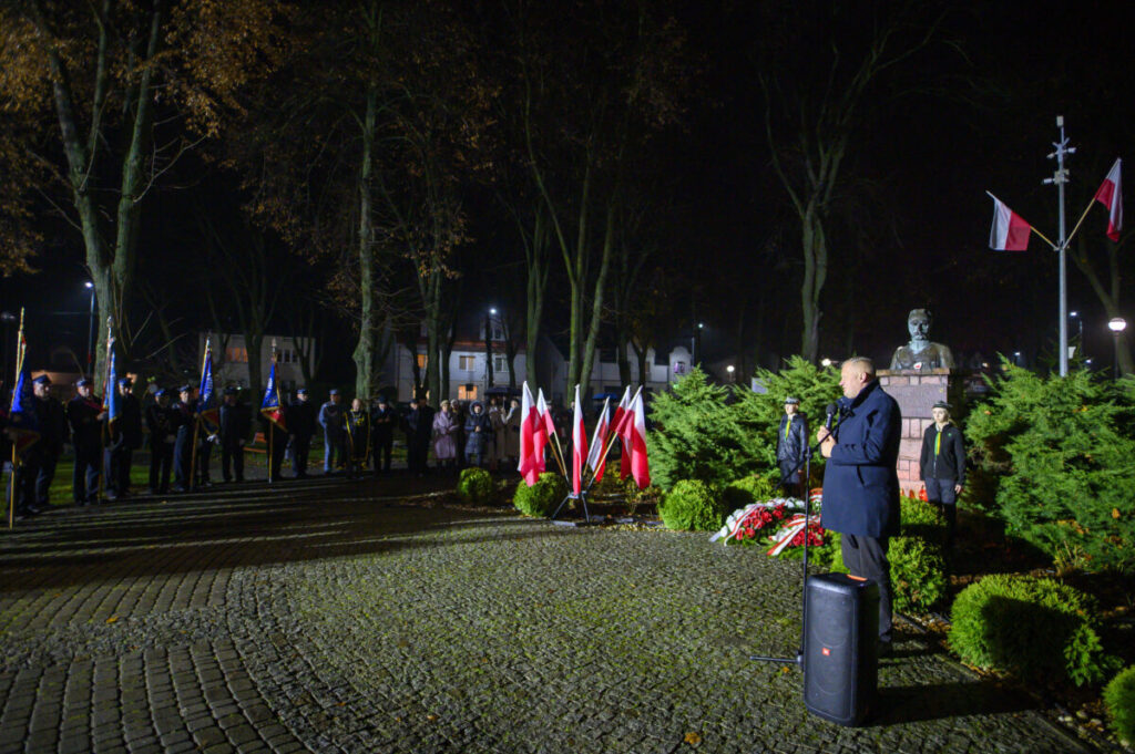 Nocne zgromadzenie na placu, z mężczyzną przemawiającym przy mikrofonie. Liczne polskie flagi na tle zielonych krzewów. Pomnik i tłum ludzi w tle.