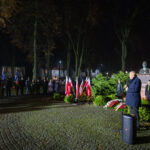 Nocne zgromadzenie na placu, z mężczyzną przemawiającym przy mikrofonie. Liczne polskie flagi na tle zielonych krzewów. Pomnik i tłum ludzi w tle.