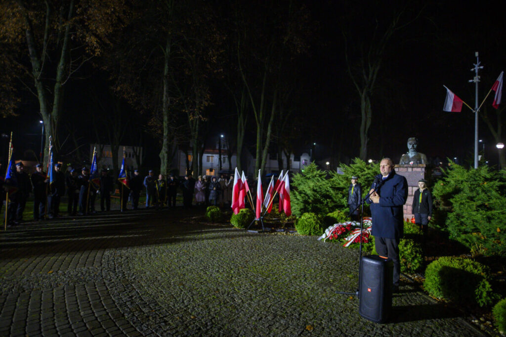 Nocna uroczystość z uczestnikami, flagami polskimi i przemawiającym. Zielone krzewy z pomnikiem w tle. Oświetlenie jasne, ciepłe.