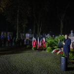 Nocna uroczystość z uczestnikami, flagami polskimi i przemawiającym. Zielone krzewy z pomnikiem w tle. Oświetlenie jasne, ciepłe.