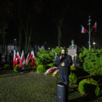 Mężczyzna przemawia nocą w parku. W tle popiersie, flagi i zielone krzewy. Ludzie zgromadzeni wokół, ciemne otoczenie.