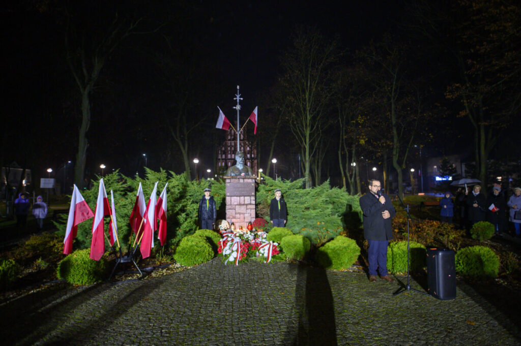 Pomnik z figurą, otoczony polskimi flagami i zielenią, w parku nocą. Przemawiający mężczyzna stoi obok mikrofonu.
