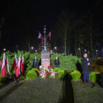 Pomnik z figurą, otoczony polskimi flagami i zielenią, w parku nocą. Przemawiający mężczyzna stoi obok mikrofonu.