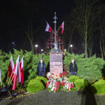 Pomnik z popiersiem, otoczony zielonymi krzewami, wieczorem. Liczne biało-czerwone flagi i kwiaty z wstążkami. Mężczyzna z mikrofonem w ciemnym płaszczu.