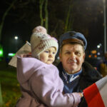 Mężczyzna w mundurze trzyma dziecko w różowym ubraniu z czapką. Dziecko trzyma polską flagę. W tle nocne rozmyte światła i drzewa.