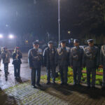 Grupa ludzi w mundurach stoi w deszczu nocą. Po lewej muzycy z instrumentami, tło ciemne z drzewami i światłami. Mundury szaro-niebieskie.