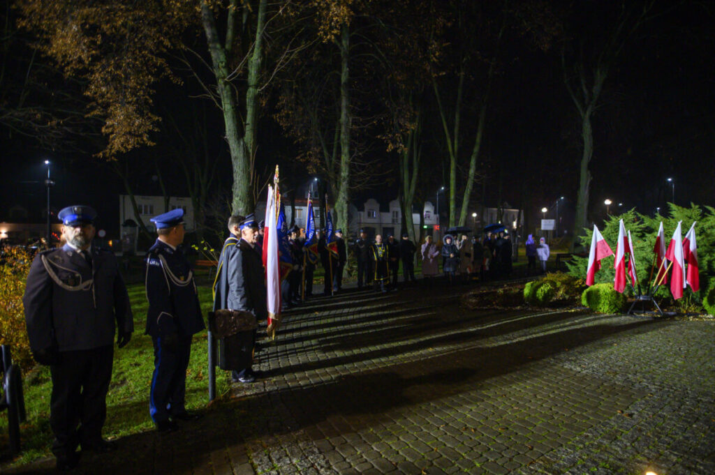 Grupa ludzi w mundurach stoi w parku nocą. Widać biało-czerwone flagi Polski oraz drzewa i bloczy w tle. Teren jest oświetlony lampami ulicznymi.