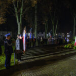 Grupa ludzi w mundurach stoi w parku nocą. Widać biało-czerwone flagi Polski oraz drzewa i bloczy w tle. Teren jest oświetlony lampami ulicznymi.