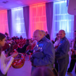 Mężczyźni w mundurach z medalami, kobieta podaje wstążki z koszyka. Jasne zasłony w tle oświetlone na niebiesko i czerwono. Salę wypełnia grupa ludzi na czerwonych krzesłach.