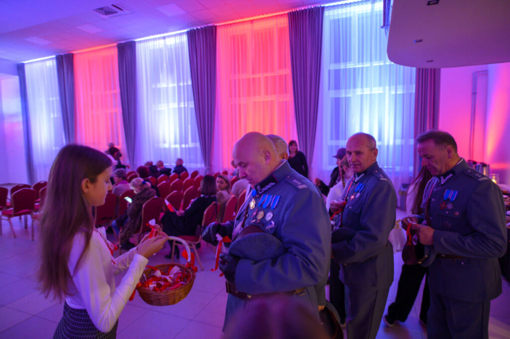 Na zdjęciu grupa osób w mundurach z medalami odbiera wstążki od dziewczyny. Tło zdobią zasłony w niebieskim i czerwonym świetle. W tle widoczne są rzędy czerwonych krzeseł i inne osoby.