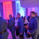 Grupa ludzi w mundurach, trzymająca kapelusze. Tło oświetlone kolorowymi światłami: niebieskim i czerwonym. Na stole po lewej widoczne dekoracje.